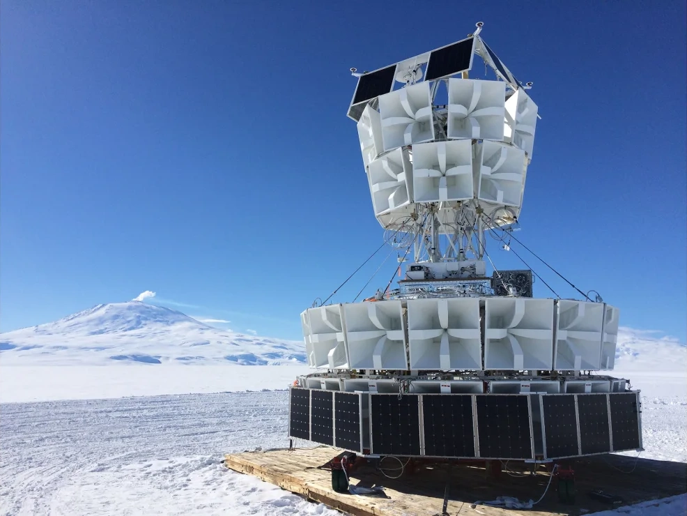 The ANITA instrument — a tall stack of radio horn antennas on a gondola with solar panels — on the Antarctic ice, a snow-capped peak behind it.