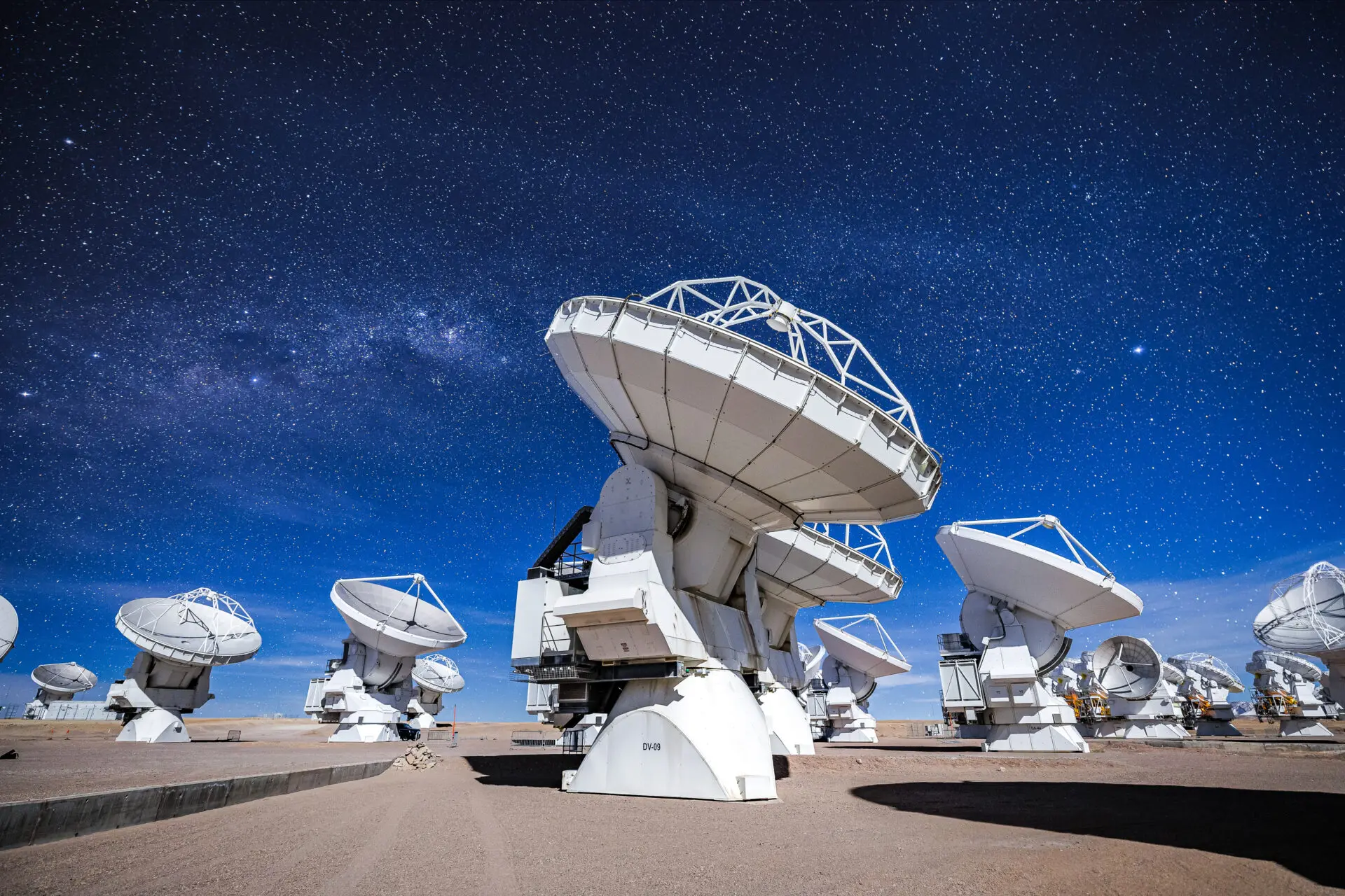 The DV-59 antenna in the foreground, with multiple ALMA antennas observing the moonlit night sky.